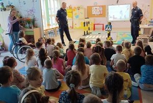 zdjęcie kolorowe: sala w przedszkolu. Dwóch Policjantów rozmawiający z dziećmi o bezpiecznym przechodzeniu przez przejście dla pieszych