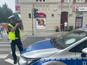 Zdjęcie kolorowe przedstawia policjanta podczas pomiaru prędkości pojazdów w ruchu drogowym.