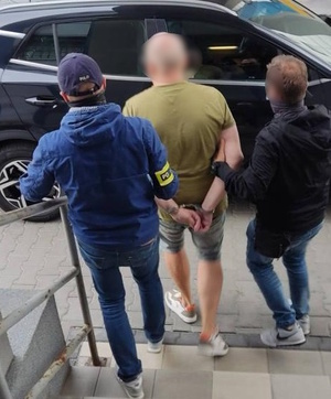 zdjęcie kolorowe: dwóch policjantów wydziału kryminalnego prowadzi mężczyznę podejrzanego o zniszczenie telefonu i naruszenie nietykalności cielesnej posła do samochodu