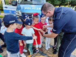 zdjęcie kolorowe: policjant zakładające na ręce przedszkolaków odblaskowe opaski