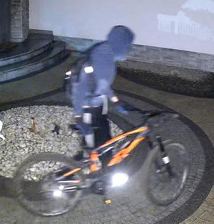 zdjęcie kolorowe: mężczyzna ubrany w ciemne spodnie, ciemna bluzę i czarną kominiarkę założoną na głowę na terenie posesji, z której ukradziono dwa rowery elektryczne