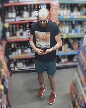 zdjęcie kolorowe: mężczyzna podejrzewany o kradzież w sklepie monopolowym, ubrany w czarną koszulkę i niebieskie spodenki, czerwone buty sportowe. Mężczyzna łysy z brodą oraz wąsem i okularami przeciwsłonecznymi, z tatuażem na lewym przedramieniu