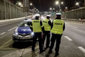 zdjęcie kolorowe: trzech policjantów drogówki stojących przy radiowozie