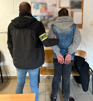 zdjęcie kolorowe: zatrzymany mężczyzna z kajdankami założonymi na ręce trzymany za ramie przez policjanta