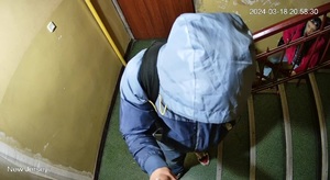 zdjęcie kolorowe: mężczyzna stojący na klatce schodowej ubrany w granatowe spodnie i niebieska kurtkę z kapturem założonym na głowę